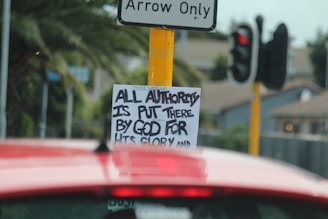A traffic sign with a sticker underneath that has handwritten text reading 'ALL AUTHORITY IS PUT THERE BY GOD FOR HIS GLORY'. A red vehicle is in the foreground, and a traffic signal and palm trees are visible in the background.