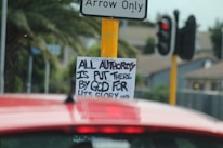A traffic sign with a sticker underneath that has handwritten text reading 'ALL AUTHORITY IS PUT THERE BY GOD FOR HIS GLORY'. A red vehicle is in the foreground, and a traffic signal and palm trees are visible in the background.