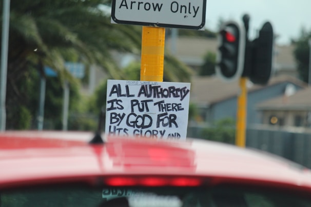 A traffic sign with a sticker underneath that has handwritten text reading 'ALL AUTHORITY IS PUT THERE BY GOD FOR HIS GLORY'. A red vehicle is in the foreground, and a traffic signal and palm trees are visible in the background.