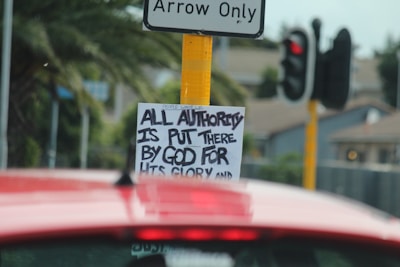 A traffic sign with a sticker underneath that has handwritten text reading 'ALL AUTHORITY IS PUT THERE BY GOD FOR HIS GLORY'. A red vehicle is in the foreground, and a traffic signal and palm trees are visible in the background.