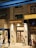 A hotel entrance at street level is brightly illuminated with a sign that reads 'Lander Grand Hotel'. Several people walk past on the sidewalk, some in casual attire. The building facade has traditional design elements with lighting fixtures flanking the entrance. Adjacent to the hotel is a storefront with a partially visible display inside.