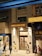 A hotel entrance at street level is brightly illuminated with a sign that reads 'Lander Grand Hotel'. Several people walk past on the sidewalk, some in casual attire. The building facade has traditional design elements with lighting fixtures flanking the entrance. Adjacent to the hotel is a storefront with a partially visible display inside.
