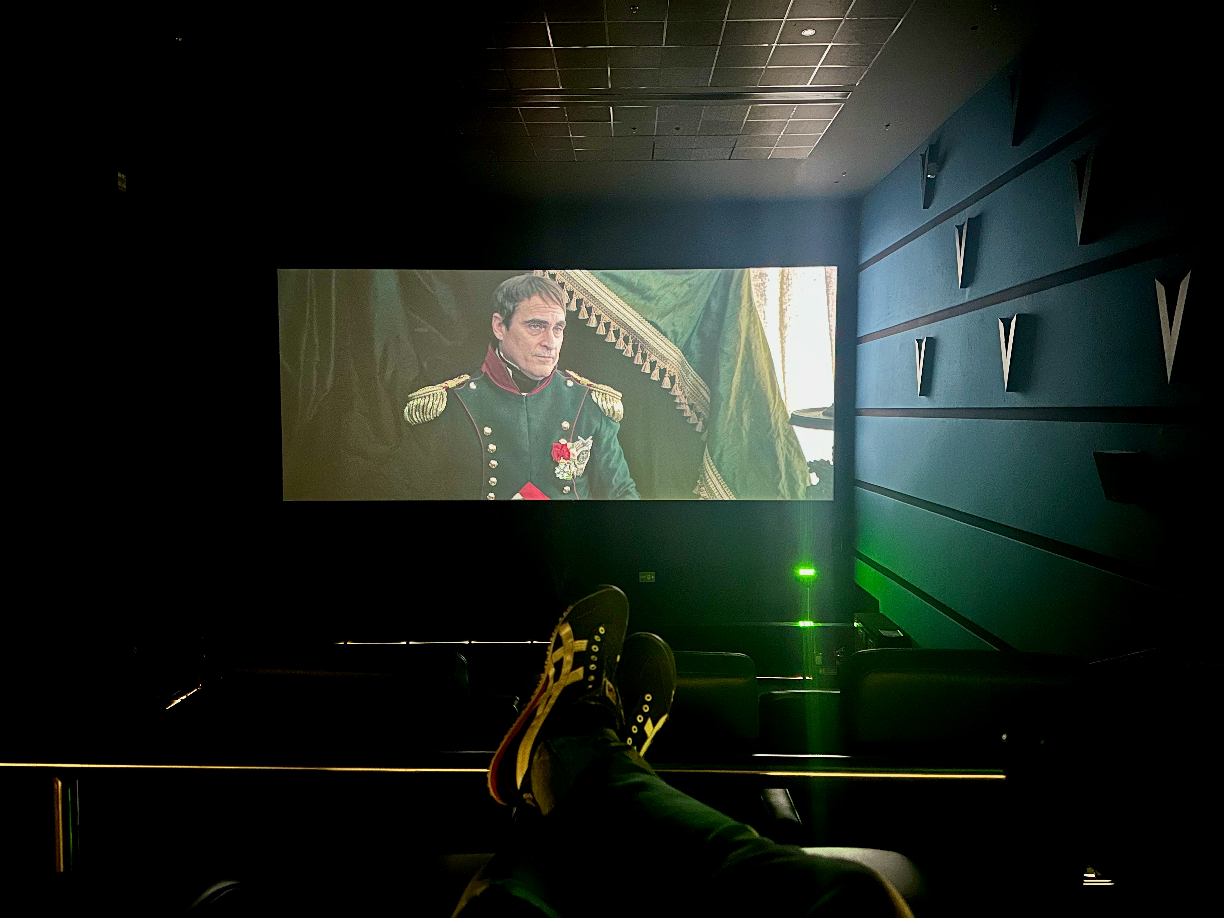 A dimly lit cinema with a large screen displays a historical film scene featuring a man in a military uniform with medals. The cinema seats are visible, and a person’s feet are resting on the back of one of the seats. The room has a modern design with structural elements on the walls.