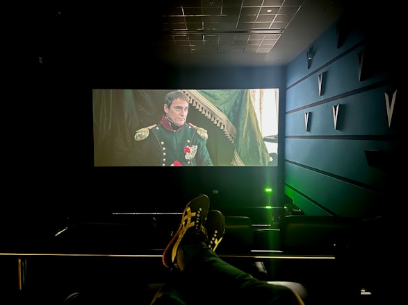 A dimly lit cinema with a large screen displays a historical film scene featuring a man in a military uniform with medals. The cinema seats are visible, and a person’s feet are resting on the back of one of the seats. The room has a modern design with structural elements on the walls.