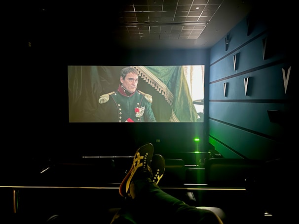 A dimly lit cinema with a large screen displays a historical film scene featuring a man in a military uniform with medals. The cinema seats are visible, and a person’s feet are resting on the back of one of the seats. The room has a modern design with structural elements on the walls.