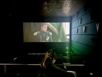 A dimly lit cinema with a large screen displays a historical film scene featuring a man in a military uniform with medals. The cinema seats are visible, and a person’s feet are resting on the back of one of the seats. The room has a modern design with structural elements on the walls.