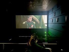 A dimly lit cinema with a large screen displays a historical film scene featuring a man in a military uniform with medals. The cinema seats are visible, and a person&rsquo;s feet are resting on the back of one of the seats. The room has a modern design with structural elements on the walls.