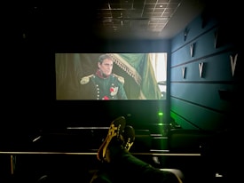 A dimly lit cinema with a large screen displays a historical film scene featuring a man in a military uniform with medals. The cinema seats are visible, and a person’s feet are resting on the back of one of the seats. The room has a modern design with structural elements on the walls.