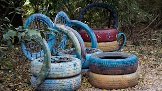 A collection of painted tires arranged creatively in an outdoor setting, surrounded by foliage. The tires are stacked and positioned in circular formations, featuring shades of blue, red, and yellow with decorative patterns.