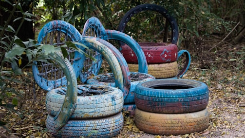 A collection of painted tires arranged creatively in an outdoor setting, surrounded by foliage. The tires are stacked and positioned in circular formations, featuring shades of blue, red, and yellow with decorative patterns.