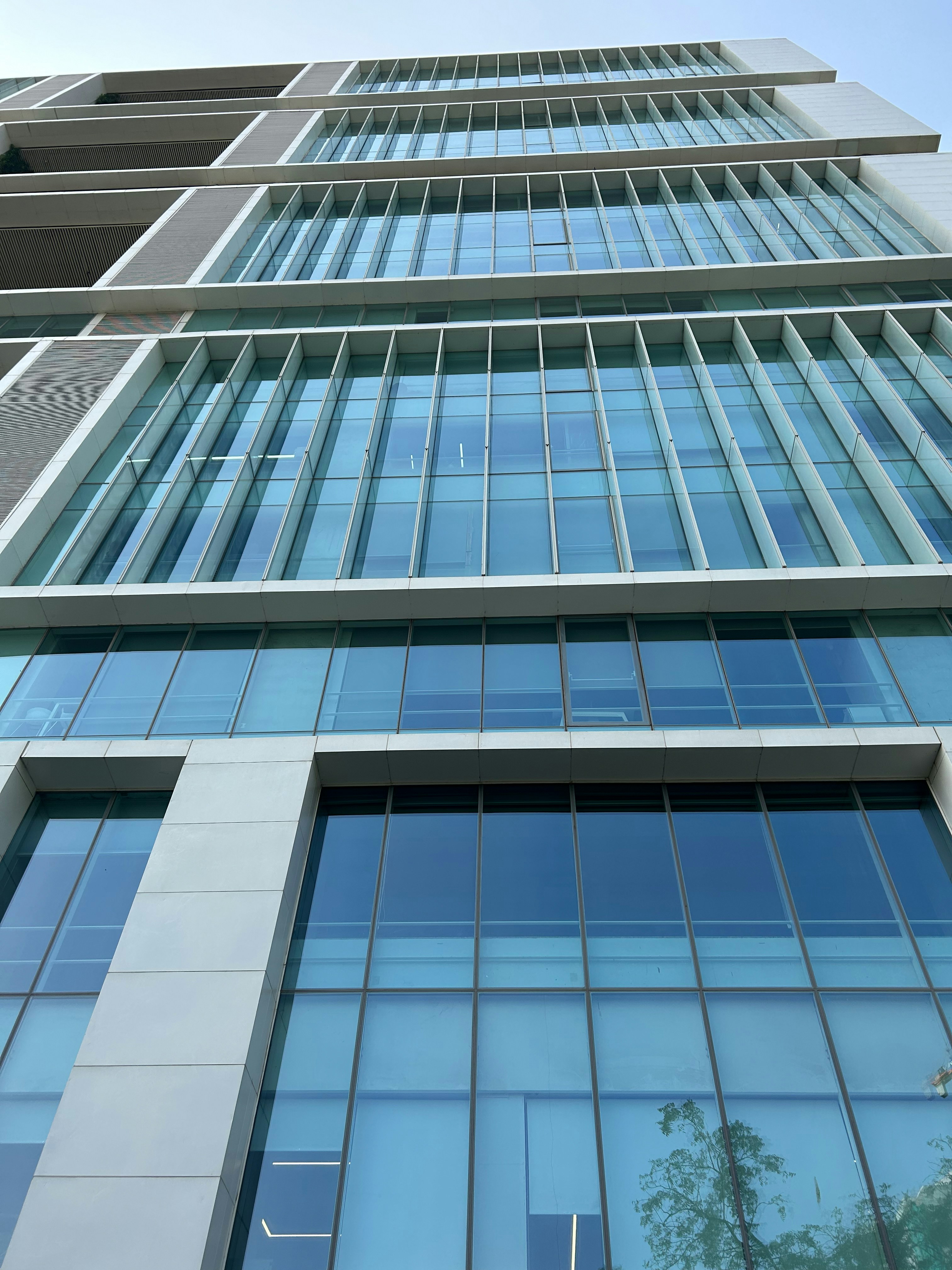 A modern architectural design showcasing a high-rise building with large, reflective glass windows. The windows are arranged in a grid-like pattern, and the building features sharp, clean lines. The sky above is clear with a hint of vegetation on the right.