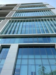 A modern architectural design showcasing a high-rise building with large, reflective glass windows. The windows are arranged in a grid-like pattern, and the building features sharp, clean lines. The sky above is clear with a hint of vegetation on the right.