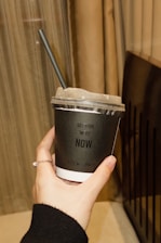 A hand holding a black disposable cup with a plastic lid and a black straw. The cup has white lettering that says 'GET YOUR 'MEDS' NOW'. The hand is adorned with a thin ring. In the background, there are beige curtains and a part of a dark-colored object, possibly a piece of furniture.