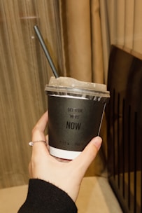 A hand holding a black disposable cup with a plastic lid and a black straw. The cup has white lettering that says 'GET YOUR 'MEDS' NOW'. The hand is adorned with a thin ring. In the background, there are beige curtains and a part of a dark-colored object, possibly a piece of furniture.
