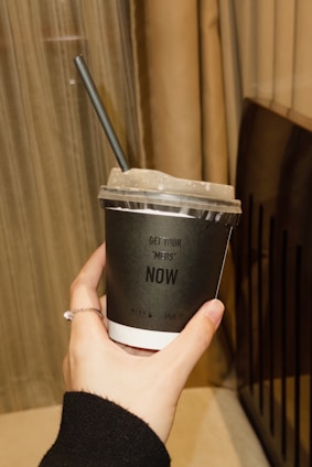 A hand holding a black disposable cup with a plastic lid and a black straw. The cup has white lettering that says 'GET YOUR 'MEDS' NOW'. The hand is adorned with a thin ring. In the background, there are beige curtains and a part of a dark-colored object, possibly a piece of furniture.