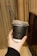 A hand holding a black disposable cup with a plastic lid and a black straw. The cup has white lettering that says 'GET YOUR 'MEDS' NOW'. The hand is adorned with a thin ring. In the background, there are beige curtains and a part of a dark-colored object, possibly a piece of furniture.