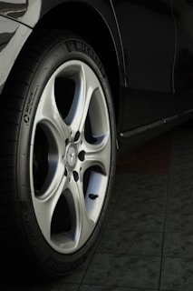 A close-up view of a car wheel with a silver alloy rim and a black tire featuring the Michelin logo. The floor beneath the car appears to be tiled.
