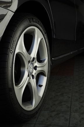A close-up view of a car wheel with a silver alloy rim and a black tire featuring the Michelin logo. The floor beneath the car appears to be tiled.