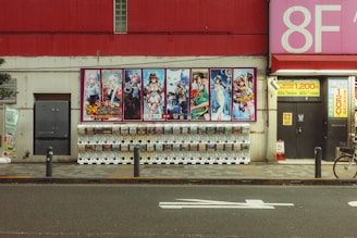 A street scene featuring a row of gachapon vending machines lined up against a wall. Above the machines, there is a large colorful poster displaying anime-style characters. The background wall is a mix of red and cream colors, with various signs and posters, including a large pink sign indicating '8F'. The sidewalk in front of the machines has a tiled pattern, and a bicycle is partially visible on the right side of the image.