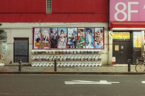 A street scene featuring a row of gachapon vending machines lined up against a wall. Above the machines, there is a large colorful poster displaying anime-style characters. The background wall is a mix of red and cream colors, with various signs and posters, including a large pink sign indicating '8F'. The sidewalk in front of the machines has a tiled pattern, and a bicycle is partially visible on the right side of the image.