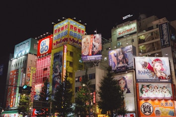 Brightly lit urban scene featuring tall buildings adorned with colorful anime and advertisement billboards. Neon lights and vibrant signage dominate the facade, creating a lively and bustling atmosphere. Several anime characters are prominently displayed on large posters.