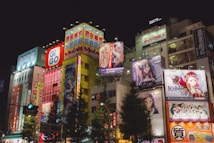 Brightly lit urban scene featuring tall buildings adorned with colorful anime and advertisement billboards. Neon lights and vibrant signage dominate the facade, creating a lively and bustling atmosphere. Several anime characters are prominently displayed on large posters.