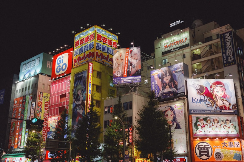 Brightly lit urban scene featuring tall buildings adorned with colorful anime and advertisement billboards. Neon lights and vibrant signage dominate the facade, creating a lively and bustling atmosphere. Several anime characters are prominently displayed on large posters.