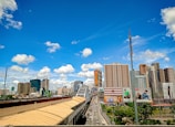 A vibrant urban landscape featuring a busy highway flanked by modern skyscrapers and large billboards. The sky is clear with a few scattered clouds, indicating a sunny day. The foreground shows a bridge over the highway, with multiple vehicles traveling in both directions.