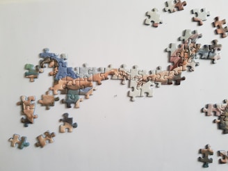 A partially completed jigsaw puzzle on a light surface, depicting fragments of a famous artwork with two hands reaching towards each other. Several loose pieces are scattered around the central image.