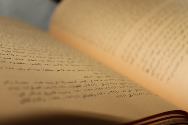 A close-up view of an open book with Arabic text. The pages are curved and the focus is on the middle of the book, with the background slightly blurred.