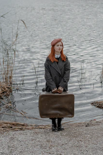 a woman with red hair is holding a suitcase by the water