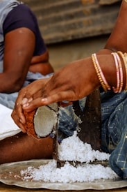 A person is grating a coconut using a manual grater over a metal tray. The hand is adorned with several bangles, and coconut shavings are accumulating on the tray. Another person is partially visible in the background.