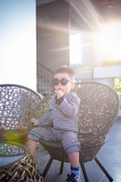 A child wearing sunglasses sits on a woven chair, dressed in a stylish checkered outfit. The setting is bright with sunlight streaming in, creating a warm and relaxed atmosphere. The child is holding something to their mouth, possibly a candy or snack.