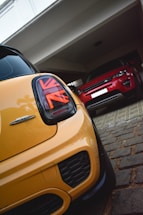 A close-up view of a yellow car with Union Jack-themed taillights parked in focus, while a red car is seen parked further inside a garage. Both vehicles are parked on a tiled and paved driveway under a white structure.