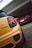 A close-up view of a yellow car with Union Jack-themed taillights parked in focus, while a red car is seen parked further inside a garage. Both vehicles are parked on a tiled and paved driveway under a white structure.