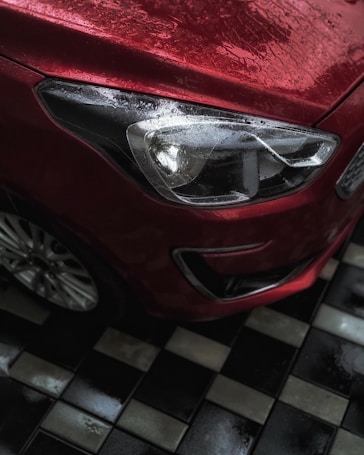 The image captures the front corner of a red car, with water droplets on its surface, indicating a recent rainfall or car wash. The car's headlight is prominently visible, reflecting light which adds to the shiny texture. The car is parked on a wet black and white checkered floor, adding contrast and interest to the scene.