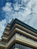 A multi-story modern building with a light beige facade and large, dark glass windows seen from a low angle. The structure is set against a backdrop of a cloudy sky with patches of blue.