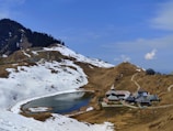 An alpine landscape with snow-covered mountains surrounding a small, serene lake. A cluster of traditional wooden buildings with steep roofs is situated near the lake, nestled in a valley. Sparse trees are visible on the hills, and the sky is clear with a few scattered clouds.