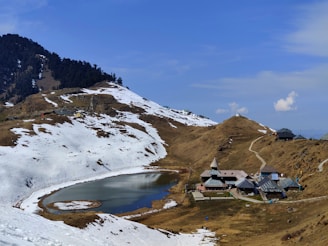 An alpine landscape with snow-covered mountains surrounding a small, serene lake. A cluster of traditional wooden buildings with steep roofs is situated near the lake, nestled in a valley. Sparse trees are visible on the hills, and the sky is clear with a few scattered clouds.