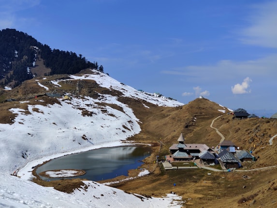 An alpine landscape with snow-covered mountains surrounding a small, serene lake. A cluster of traditional wooden buildings with steep roofs is situated near the lake, nestled in a valley. Sparse trees are visible on the hills, and the sky is clear with a few scattered clouds.