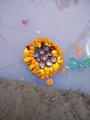 A circular arrangement of marigold flowers in orange and yellow colors floats on water. Within the floral circle, small clay lamps with lit candles are visible. The reflection of a structure or people is slightly seen in the water, and there are patches of green leaves scattered around. The setting appears to be close to the edge of a muddy area.