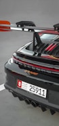 A close-up of the rear section of a sleek sports car featuring a prominent spoiler and a UAE license plate. The car's glossy black finish is highlighted with red accents, and the rear lights are illuminated.