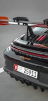 A close-up of the rear section of a sleek sports car featuring a prominent spoiler and a UAE license plate. The car's glossy black finish is highlighted with red accents, and the rear lights are illuminated.