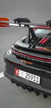 A close-up of the rear section of a sleek sports car featuring a prominent spoiler and a UAE license plate. The car's glossy black finish is highlighted with red accents, and the rear lights are illuminated.