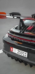 A close-up of the rear section of a sleek sports car featuring a prominent spoiler and a UAE license plate. The car's glossy black finish is highlighted with red accents, and the rear lights are illuminated.