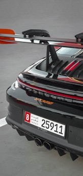 A close-up of the rear section of a sleek sports car featuring a prominent spoiler and a UAE license plate. The car's glossy black finish is highlighted with red accents, and the rear lights are illuminated.