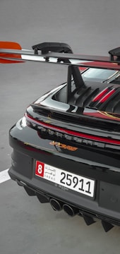 A close-up of the rear section of a sleek sports car featuring a prominent spoiler and a UAE license plate. The car's glossy black finish is highlighted with red accents, and the rear lights are illuminated.