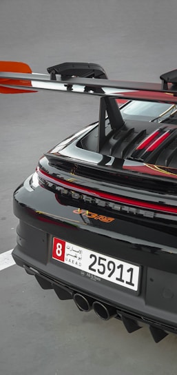 A close-up of the rear section of a sleek sports car featuring a prominent spoiler and a UAE license plate. The car's glossy black finish is highlighted with red accents, and the rear lights are illuminated.