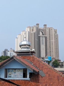 A foreground features a mosque with a red-tiled roof and a silver dome atop its roof. In the background, several tall modern high-rise buildings dominate the skyline. There is a clear blue sky above, and some greenery is visible near the buildings.