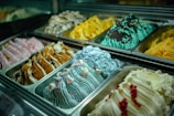 Rows of colorful gelato flavors are displayed in metal trays. Each flavor has a unique texture and color, with decorative toppings like berries, marshmallows, and sprinkles. The lighting highlights the vibrant colors and creamy texture of the gelato.